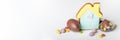 Broken Easter chocolate eggs, house shaped cookie and colorful decorations on a light background. Banner. Easter