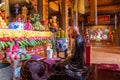 Bronze statue of a monk kneeling in front of richly decorated altar with harvested food inside a Buddhist temple