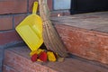 Broom with a red handle and a yellow dustpan, propped against a brick wall next to a pile of autumn leaves on a wooden brown porch