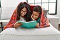 Brother and sister covering with blanket reading book lying on sofa at home