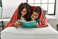 Brother and sister covering with blanket reading book lying on sofa at home