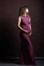 On brown background is a young pregnant woman, a full-length photograph, looking to the side, beautiful makeup and hairstyle, a
