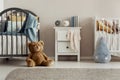 Brown teddy bear on the wooden floor of scandinavian baby bedroom interior with white nightstand and cribs