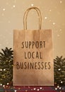 Brown Christmas paper bag with `support local businesses` message in front of a beige background with pine cone props