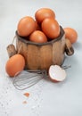 Brown eggs in small wooden bucket with yolk and shell on light kitchen table with metal whisk