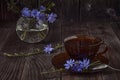A brown glass cup with chicory stands on a brown wooden table. On a saucer branch with chicory flowers