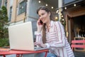 Brown-haired female working at laptop in open air cafe, talking on phone Royalty Free Stock Photo