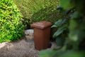 Brown large plastic bin for trash on backyard, around of tree hedge