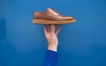 A brown leather shoe with a textured upper and a creamcolored sole is being held by a hand against a solid blue background Royalty Free Stock Photo