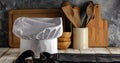 White chef`s hat and wooden table background with dark wall and kitchen utensils.