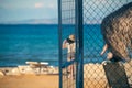 Brown Sparrow Sits On Gray Metal Fence Against Background Of Beach With Vacationers, Blue Ionian Sea, Balkan Mountains And Sky Royalty Free Stock Photo