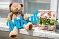 Brown Teddy bear sitting on the windowsill with a blue sign in his paws, next to two pots of flowers. Royalty Free Stock Photo
