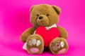 Brown toy cartoon teddy bear sits on a pink background with red paper heart