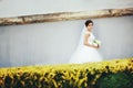 Brunette bride walks along a white wall behind yellow bushes
