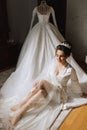 A brunette bride wearing a satin robe sits on the floor next to her dress.