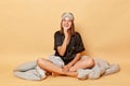 Brunette Caucasian young girl sitting floor with blanket wearing black pajama and sleep eye mask isolated over beige background Royalty Free Stock Photo