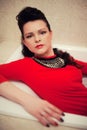 Brunette with curly hair in a red dress lies in an empty white bathtub Royalty Free Stock Photo