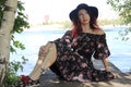 Brunette with curly red curls in a black hat and summer dress, sits on a wooden bridge on the riverbank Royalty Free Stock Photo