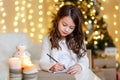 A brunette gilr in front of fur-tree writing a letter to santa claus. A girl dreaming. New year`s eve. Christmas