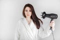 Brunette girl in bathrobe with hair dryer dries her hair against a white background. Royalty Free Stock Photo