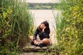 A brunette girl in a black jacket sits on a wooden bridge on the bank of a pond, propping her head up with her hand Royalty Free Stock Photo