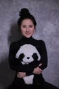 Portrait of a brunette girl in a black turtleneck holding big fluffy panda-toy against a gray wall