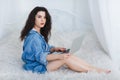 Brunette girl in denim shirt, sits on the bed, and uses a modern tablet. Indoors in the bedroom. Royalty Free Stock Photo