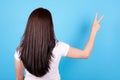 Brunette girl with long hair showing peace gesture, view from behind. Isolated on blue background