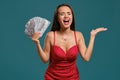 Brunette girl with a long hair, wearing a sexy red dress is posing holding a fan of hundred dollar bills against a blue Royalty Free Stock Photo