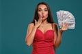 Brunette girl with a long hair, wearing a sexy red dress is posing holding a fan of hundred dollar bills against a blue Royalty Free Stock Photo