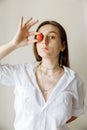 A brunette girl looks at the viewer. With one hand, the girl covers her eye with a red cherry tomato.