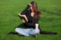 A brunette girl sits in a green meadow and indignantly reads a hardback book. Royalty Free Stock Photo