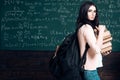 Brunette girl standing with heavy pile of books and big black backpack in front of green board full of writing Royalty Free Stock Photo