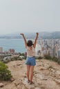 Brunette girl stands on a cliff of a high mountain with arms raised up and looks at the panorama of the city