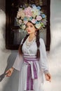 Brunette girl in a white ukrainian authentic national costume and a wreath of flowers is posing against a white hut