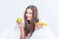 The brunette is having breakfast with fruit. The girl sits on the bed wrapped in a blanket and holds a banana and an apple Royalty Free Stock Photo