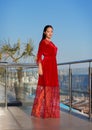 A brunette lady standing on a balcony on a blue sky background. A fashionable woman in a long red dress. A female on a Royalty Free Stock Photo