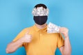 Brunette man in protection mask, paper money on the face, shows thumbs up. Isolated on blue background