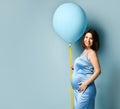 Pregnant female in blue dress. Smiling, put hand on tummy, holding balloon by yellow ribbon, posing sideways on blue background Royalty Free Stock Photo