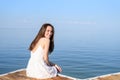 Brunette sitting on the pier in a white sundress, smiling, looking at the camera in half a turn on the background of the sea, rear Royalty Free Stock Photo