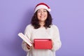 Brunette smiling woman in santa hat with opened gift box, looks happy at camera, has great present, being in good mood, standing Royalty Free Stock Photo