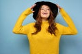 A brunette stands on a blue background in a yellow sweater and smiles with head thrown back and the edges of the black
