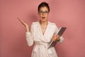 A brunette stands on a pink background with collected hair holding a laptop, opening his mouth in surprise, looks into