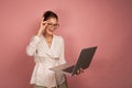 A brunette stands on a pink background with her hair collected and happily looks into the laptop while adjusting her