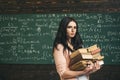 Brunette student walking with two heaps of books in front of green board full of writing. Math undergraduate preparing