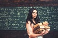 Brunette student walking with two heaps of books in front of green board full of writing. Math undergraduate preparing Royalty Free Stock Photo