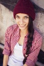 Brunette teenage girl in hipster outfit (jeans shorts, keds, plaid shirt, hat) with a skateboard at the park