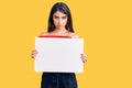 Brunette teenager girl holding blank empty banner thinking attitude and sober expression looking self confident