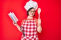 Brunette woman with down syndrome wearing professional baker apron reading cooking recipe book winking looking at the camera with Royalty Free Stock Photo