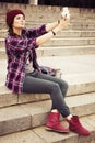 Brunette woman in hipster outfit sitting on steps and taking selfie on retro camera on the street. Toned image Royalty Free Stock Photo